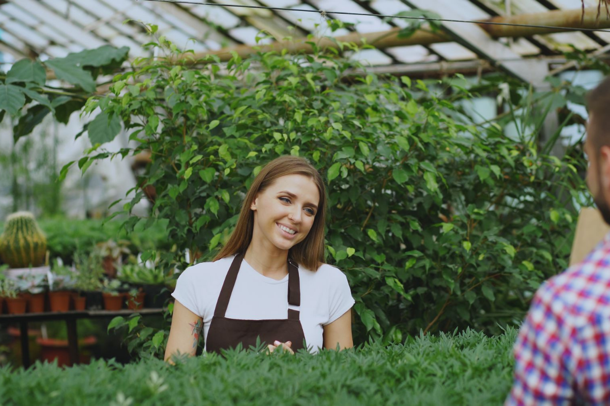 Nursery staff member talking with a customer among potted plants in a greenhouse